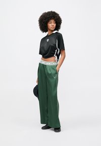 Νεαρή γυναίκα με σγουρά μαλλιά, φορώντας μαύρο κοντό αθλητικό τοπ και πράσινα φαρδιά παντελόνια Nike, κρατώντας μαύρο καπέλο, στέκεται σε λευκό φόντο.