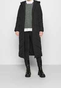 Black long puffer coat with hood, quilted pattern, paired with a grey sweater and white shirt, black jeans, and chunky black boots.