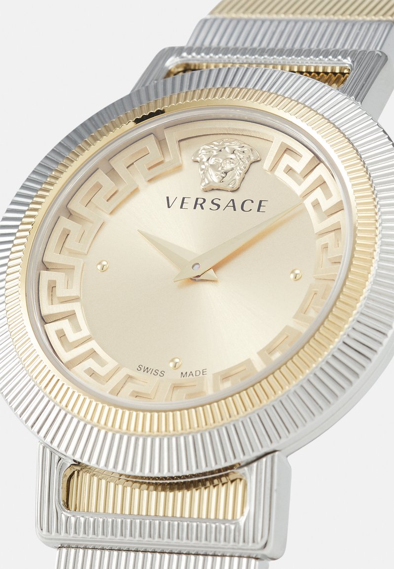 Versace-Uhr mit einem runden Gehäuse aus Edelstahl in Gold und Silber, strukturiertem Lünette, goldenem Zifferblatt und griechischem Schlüsselmotiv um das Ziffernblatt.