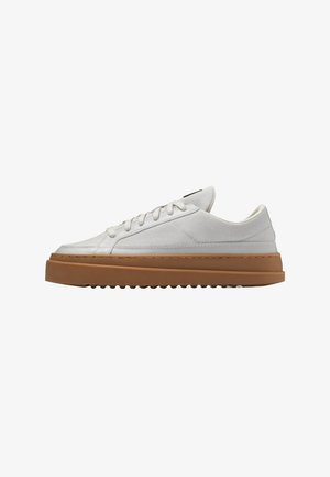 White low-top sneaker with canvas upper, white laces, thick brown rubber sole, and ridged tread on a plain white background.