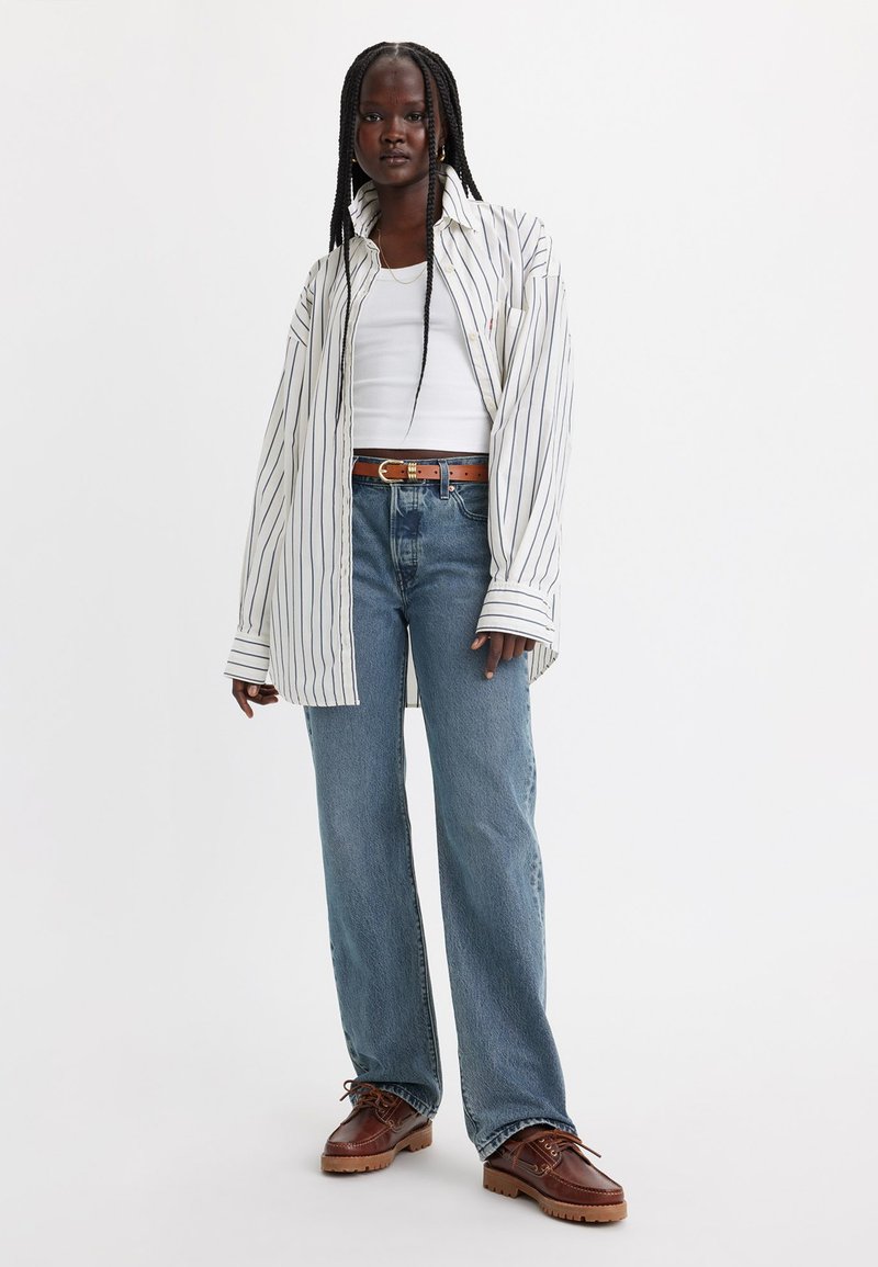 Wit gestreepte oversized shirt, witte tanktop, blauwe rechte jeans, bruine schoenen met gestructureerde zolen, en een bruine leren riem.