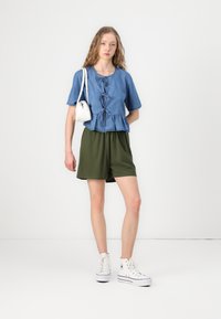 Denim majica s kratkimi rokavi z vrvico in peplum robom, v kombinaciji z olivno zelenimi kratkimi hlačami in belimi visokimi supergami. Dodatka: mala bela torbica.