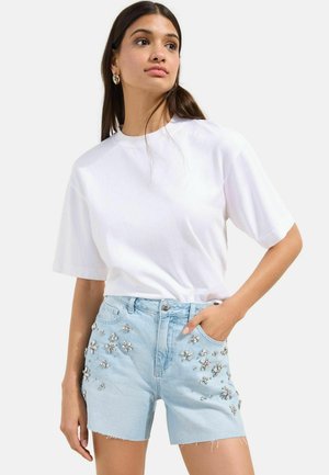Žena nosí biele tričko s krátkym rukávom a svetlomodré denimové šortky so striebornými kvetinovými ozdobami, pózuje s jednou rukou v vrecku.