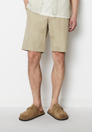 Beige shorts featuring a relaxed fit and mid-thigh length, made from lightweight fabric. Paired with brown suede slip-on shoes.
