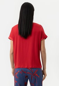 Röd t-shirt med korta ärmar, avslappnad passform, slät textur och uppvikta ärmslut. Bakifrån visas med flödande svart hår och mönstrade blå byxor.