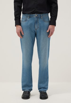 Person trägt hellblaue Jeans mit geradem Bein, schwarzes Langarmshirt und schwarze Loafers und steht vor einfachem Hintergrund.