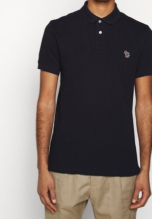 Homme portant un polo noir avec un petit logo zèbre multicolore brodé sur la poitrine et un pantalon beige.