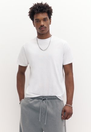 Nuori mies kiharat hiukset, päällään yksinkertainen valkoinen t-paita, harmaat nyörilliset housut, hopeinen ketjukaulakoru ja rannekoru, seisoo valkoisen taustan edessä.