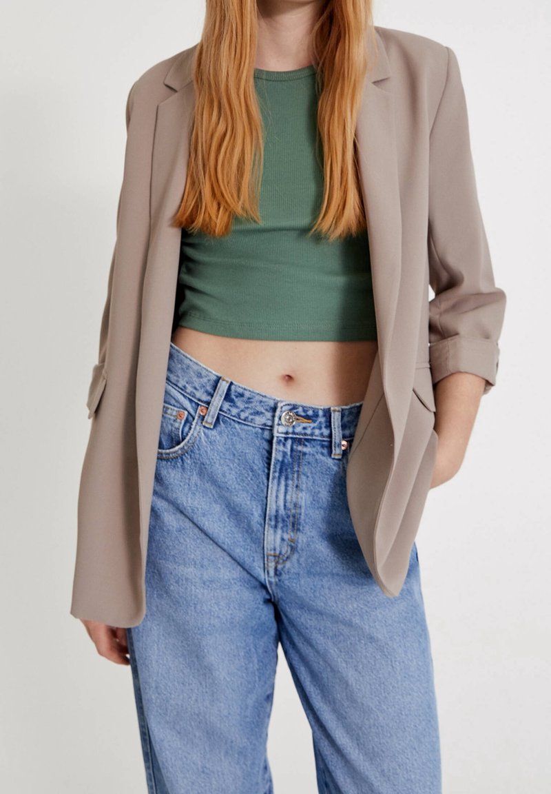 Personne portant un blazer beige par-dessus un haut court vert et un jean bleu clair taille haute, avec une main dans la poche, sur un fond blanc.