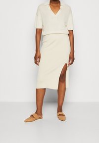 Un mannequin de mode portant un haut en tricot crème et une jupe assortie avec une fente sur le côté, accompagnés de mules beiges, sur un fond uni.