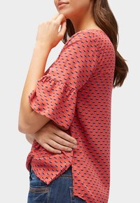 Punainen pusero, jossa on lyhyet röyhelöiset hihat ja pieni kukkakuvio. Kangas on sileää ja helmassa on sivuhalkiot.