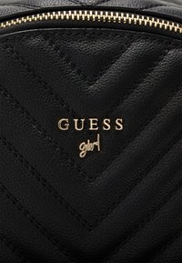 Čierna prešívaná taška z umlej kože so zlatým zipsom a logom, na ktorom je nápis "GUESS girl." Povrch má štruktúrovaný chevron vzor.
