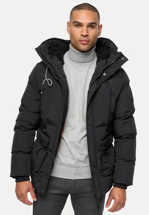 Schwarze Steppjacke mit Kapuze, versehen mit einem gesteppte Design, Fronttaschen und einem Reißverschluss. Getragen über einem grauen Rollkragensweater.