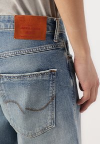 Jean en denim bleu clair avec une finition délavée, portant une étiquette en cuir marron avec l'inscription "JACK & JONES ARCHIVE" près de la poche arrière.