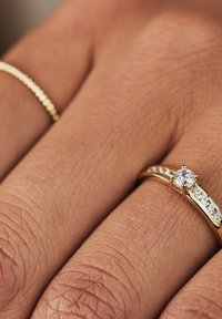 Goldring mit einem zentralen runden Diamanten, akzentuiert von kleineren Diamanten entlang des Bandes, vor einem strukturierten Hauthintergrund.