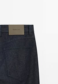 Tmavě modré džínové kalhoty s koženou nášivkou a názvem značky, zadní kapsou a čistými liniemi, které ukazují na klasický design.