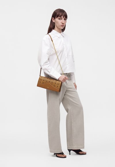 Femme portant une chemise blanche, un pantalon beige à jambes larges, des sandales noires à talons, tenant un sac à bandoulière marron à motifs avec une sangle chaîne dorée.