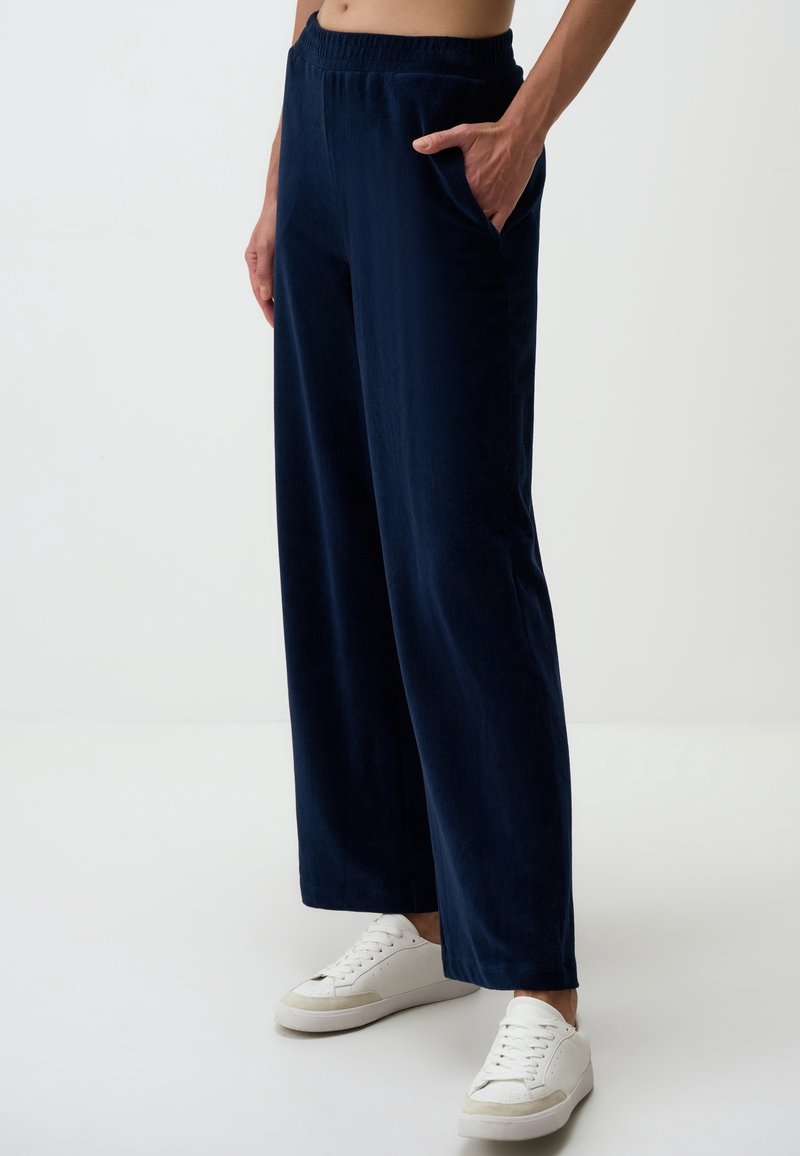 Personne portant un pantalon large bleu marine avec taille élastique et des baskets blanches, debout avec une main dans la poche sur un fond uni.