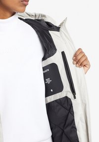 Valkoinen eristetty takki mustilla yksityiskohdilla, jossa on vetoketjullinen tasku ja etiketti sisäpuolella. Takissa on tikattu rakenne.