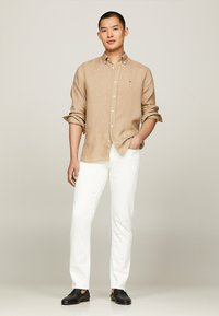 Camicia in lino color marrone chiaro con maniche arrotolate, abbinata a pantaloni bianchi slim-fit e scarpe nere slip-on, con un sottile dettaglio del logo.