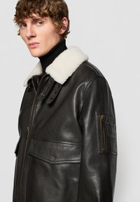 Black leather jacket featuring a white shearling collar, zipper closure, and side pockets with a branded accent on the sleeve.