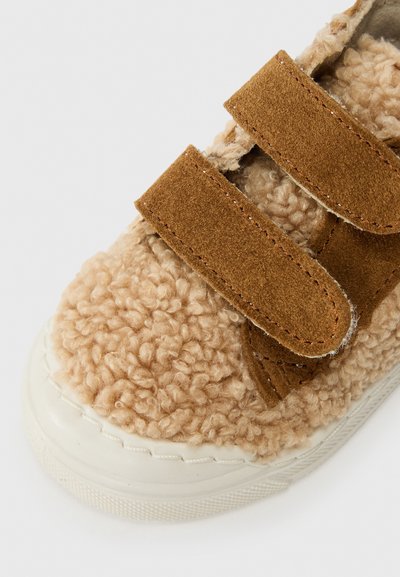 Beige, fluffy sko med teksturert stoff, brune semskede stropper og en hvit gummisåle med teksturert bunn. Glatte sømdetaljer er synlige.