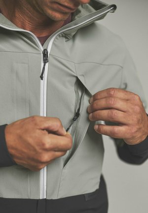 Man adjusting zipper on chest pocket of light gray jacket with white front zipper against neutral background.