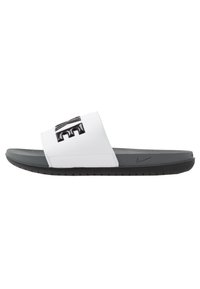 Nike šlepetės su baltu sintetiniu dirželiu, kuriame yra iškilęs juodas logotipas. Tamsiai pilka paminkštinta padas su tekstūruoto paviršiaus, užtikrinančiu sukibimą.