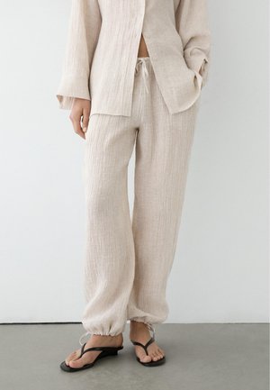 Osoba nosząca luźne, jasnobeżowe spodnie o fakturze, ze ściągaczami na sznurki, pasującą zapinaną na guziki koszulę oraz czarne sandały typu open-toe na szarej podłodze.