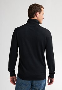 Schwarzer Strickpullover mit hohem Kragen und langen Ärmeln, der eine glatte Textur aufweist. Getragen über blauen Jeans.