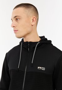 Jonge man met kort haar, gekleed in een zwarte rits hoodie met gepatternde trekkoorden en een "PROTEST" logo op de borst, kijkend naar links.