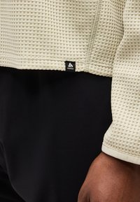 Μπεζ μακρυμάνικη μπλούζα με υφή και ετικέτα της μάρκας Odlo, φορεμένη πάνω από μαύρο παντελόνι, εμφανίζοντας μέρος του μανικιού και ένα χέρι.