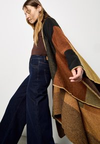 Multikleurige gebreide poncho met bruine, zwarte en mosterdkleurige blokken, franjes aan de randen, gedragen over een aansluitende bruine top en donkere wijde jeans.