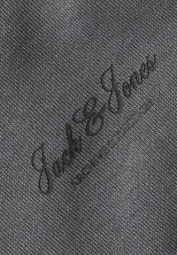 Tissu gris avec texte noir brodé « Jack & Jones » en cursive et texte en lettres plus petites en dessous.