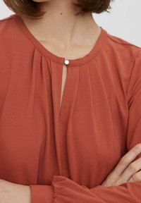 Blouse de couleur rouille avec un col rond, détails plissés à l'avant et une fermeture à bouton décoratif. Manches longues avec accents froncés.