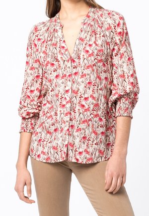 Nainen, jolla on beigehousut ja pitkähihainen kukkakuosinen paita, jossa on punaisia ja vaaleanpunaisia kukkia sekä kerätyt hihansuut, seisoo valkoista taustaa vasten.