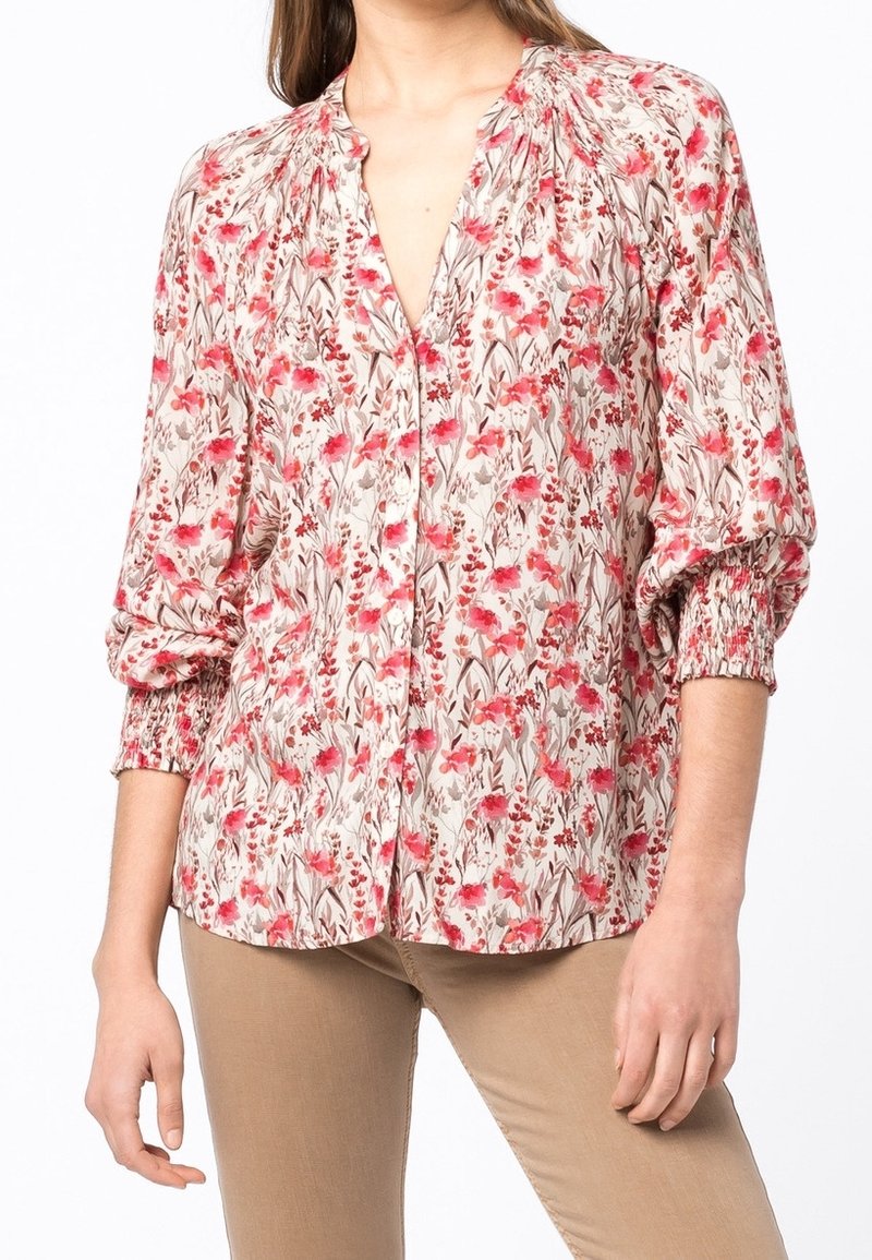 Femme portant un pantalon beige et un chemisier à manches longues à fleurs rouges et roses avec des poignets froncés, debout devant un fond blanc.