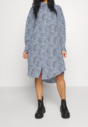 Kvinde iført en løs, lyseblå, langærmet kjole med knapper og sorte pletter samt sorte ankelstøvler, stående foran en ensfarvet baggrund.