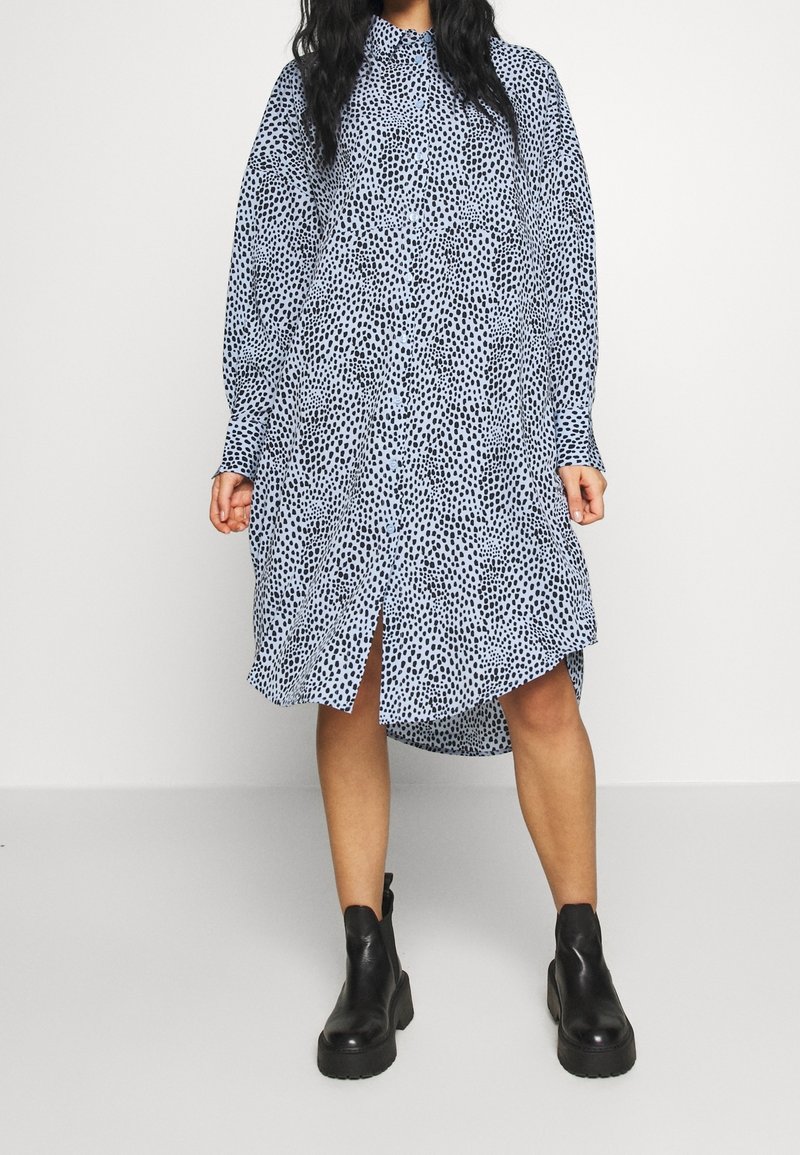 Femme portant une robe ample à manches longues, boutonnée, bleu clair avec des taches noires, ainsi que des bottines noires, debout devant un fond uni.