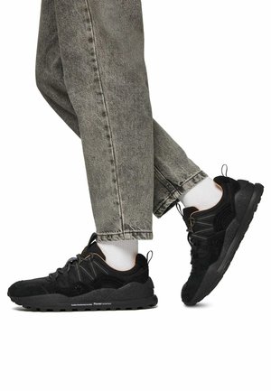Scarpe sportive nere con materiali in suede e rete, dotate di una suola testurizzata, accentuazioni a contrasto e lacci riflettenti. Indossate con jeans grigi.