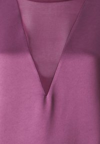 Top a scollo a V in una ricca tonalità prugna, con una texture liscia e un pannello traslucido sovrapposto al collo, che arricchisce il suo design.