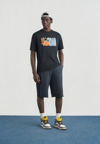 Μαύρο μπλουζάκι με πολύχρωφο γραφικό "AIR JORDAN", συνδυασμένο με σκούρο μπλε σορτς και πολύχρωμα ψηλά αθλητικά παπούτσια. Στέκεται σε μπλε χαλί.