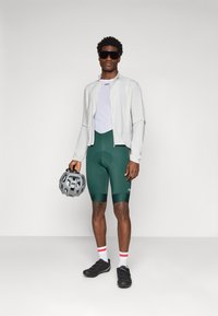 Tenue de cyclisme comprenant une veste gris clair, un haut en maille blanc, un short rembourré vert, des chaussures noires et un casque gris. Chaussettes rayées rouges et blanches.