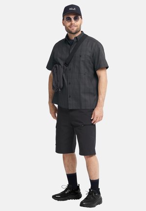 Homme portant une chemise à manches courtes à carreaux noirs, un short noir, des baskets noires, une casquette, des lunettes de soleil rondes et un sac bandoulière noir, debout sur fond blanc.