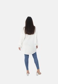Witte oversized brei trui met lange mouwen, gecombineerd met strakke blauwe jeans en doorzichtige hoge hakken, gezien van achteren.