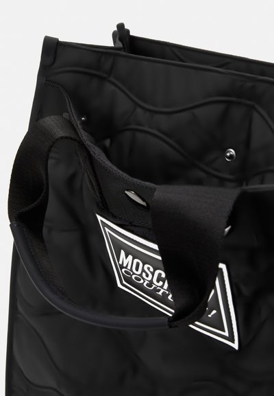 Borsa nera trapuntata realizzata in tessuto robusto, con chiusure a scatto doppie e un logo bianco prominente su una toppa triangolare. Manici lunghi neri.