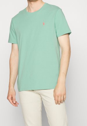 Homme portant un t-shirt à manches courtes vert menthe avec un petit logo rouge, associé à un pantalon blanc cassé, debout devant un fond uni.