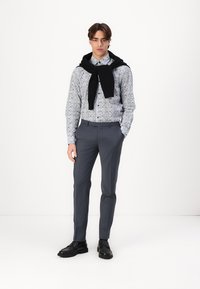 Lichtblauwe bloempatronen overhemd met knopen, antracietgrijze broek, zwarte trui over de schouders gedrapeerd, zwarte schoenen, staand voor een witte achtergrond.