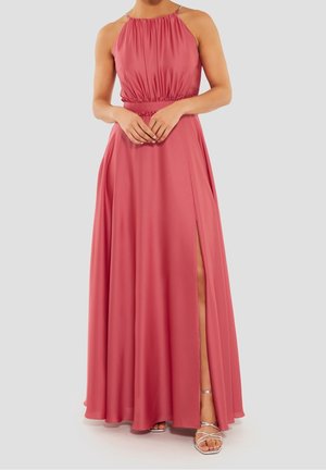 Kobieta w bezrękawnym, długim do ziemi, pudrowo-różowym sukience z wysokim kołnierzem i bocznym rozcięciem, stojąca z rękami złożonymi, widoczne srebrne sandały na paski.