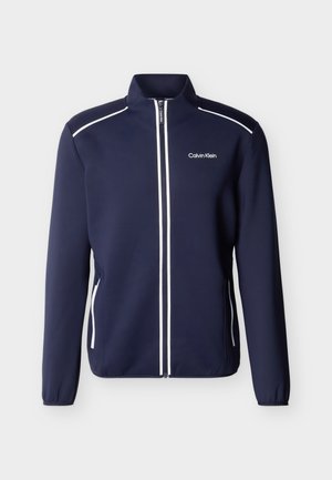 Temno modra jopica Calvin Klein z zadrgo, z belo obrobo vzdolž zadrge, ramen in žepov, z visokim ovratnikom in dolgimi rokavi.